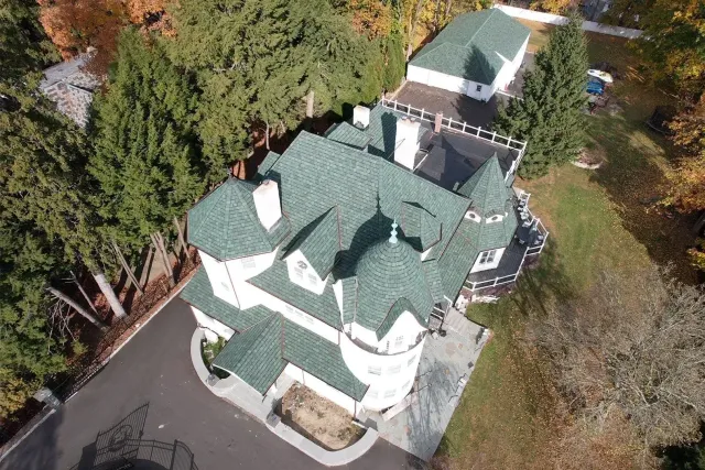 Aerial view of a white house with a green roof surrounded by trees and a driveway.