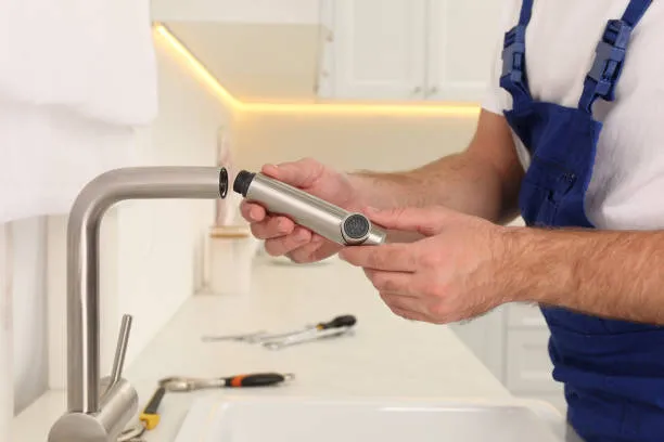 Plumber in blue overalls holding a stainless steel faucet spray head while installing a kitchen faucet