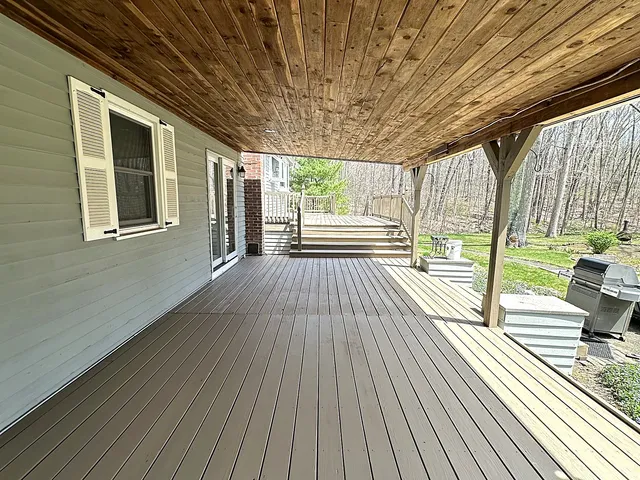 Covered rear deck in East Hampton Connecticut with freshly stained taupe brown deck boards, a natural cedar tongue-and-groove porch ceiling, timber posts, and steps down to an open second deck level facing the woods