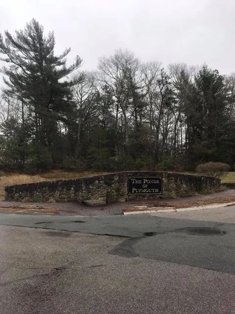 A stone sign reading The Woods of Plymouth sits at a road intersection in front of a dense forest on an overcast day.