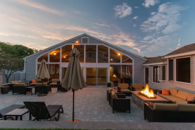 Outdoor patio with seating and a firepit, house with large glass windows, evening sky with clouds.