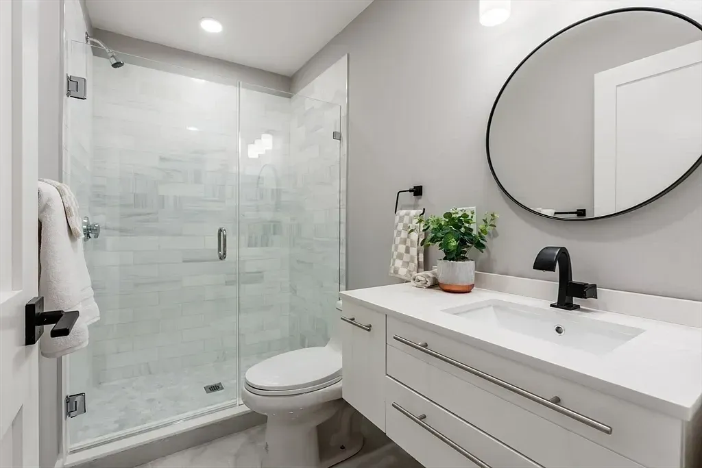 Modern bathroom with walk-in glass shower, white vanity with black faucet, circular mirror, and a potted plant on the countertop.