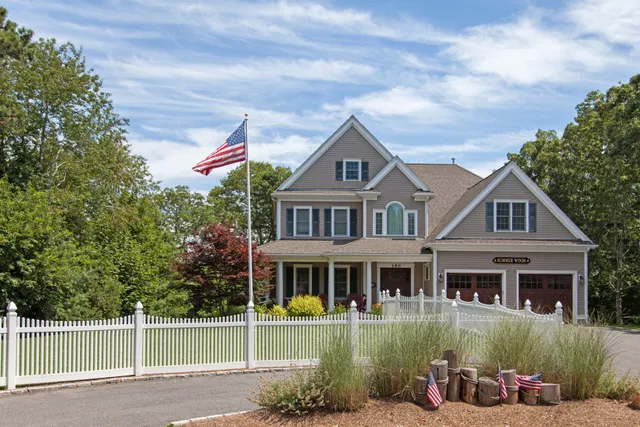 A large two-story house with beige siding and a three-car garage sits behind a white picket fence. An American flag is on a pole in the front yard. Trees and bushes surround the property under a partly cloudy sky.