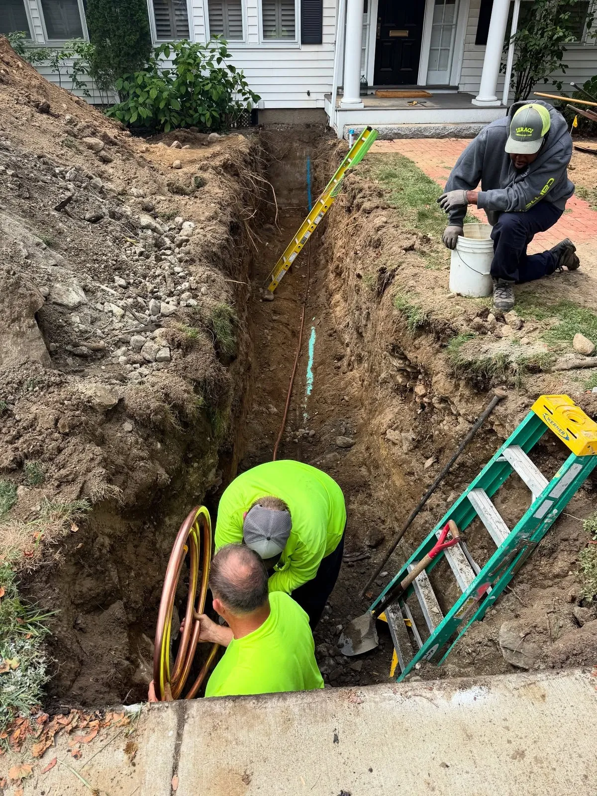 Workers installing pipes in a trench next to a house. One kneels and another holds a coil.