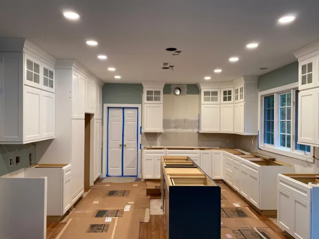 Bright unfinished kitchen with white cabinets, blue island, recessed lights, and taped flooring under renovation