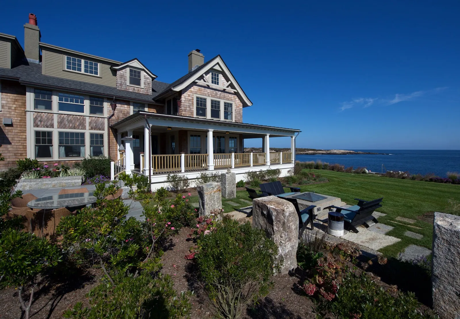 Large house with wooden shingles and a wrap-around porch, overlooking the ocean with clear blue sky, landscaped yard, patio with outdoor chairs and a fire pit.