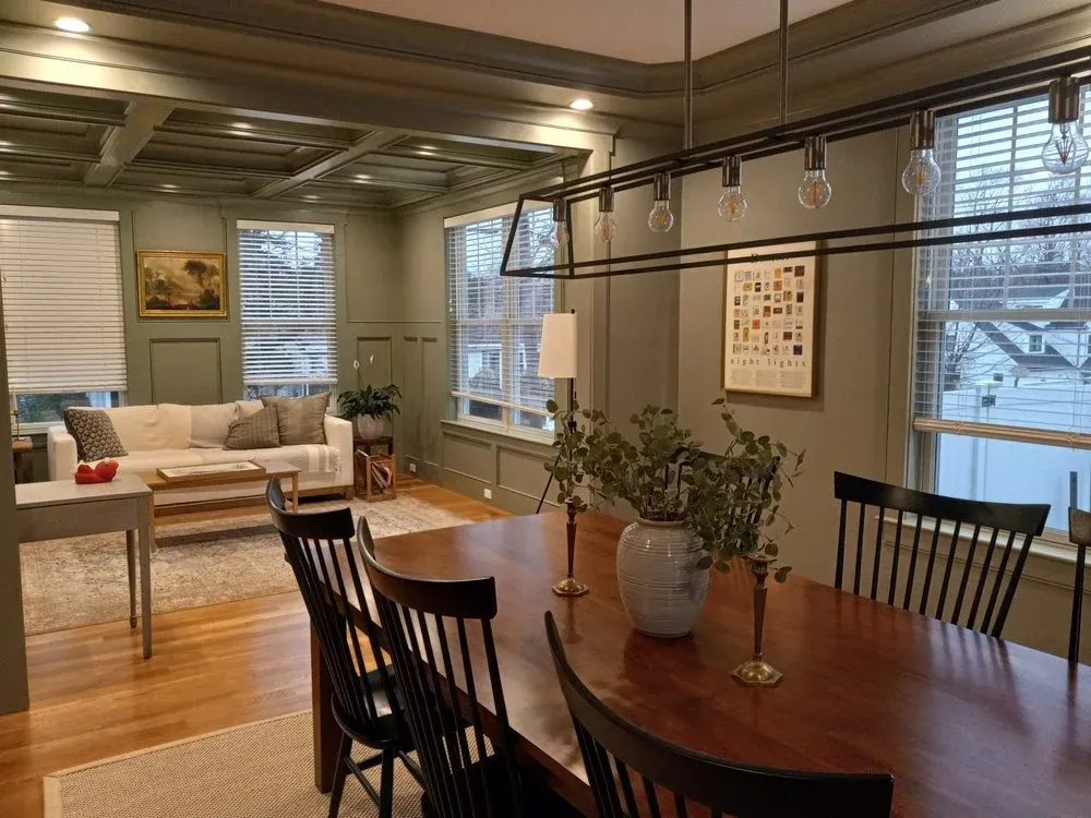 A cozy living room and dining area with green walls, wooden flooring, a large window with blinds, a white sofa with cushions, a wooden coffee table, a side table with a plant, framed artwork, a chandelier with exposed bulbs, a wooden dining table wit