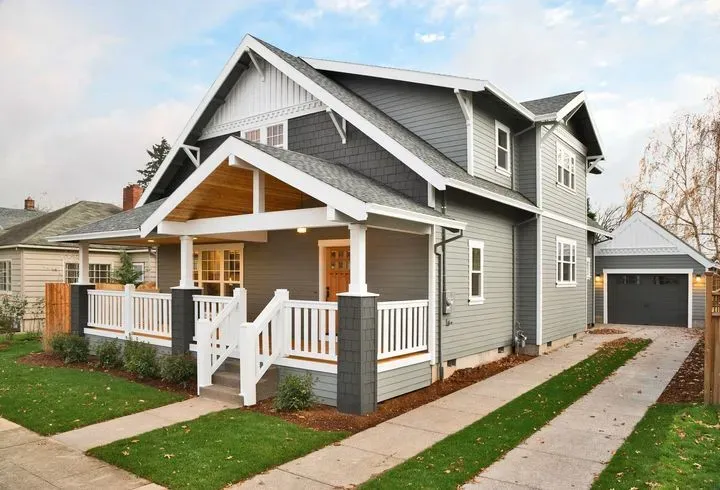 A grey, two-story craftsman-style house with a front porch, white railings, and a detached garage with a driveway.