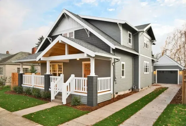 A grey, two-story craftsman-style house with a front porch, white railings, and a detached garage with a driveway.