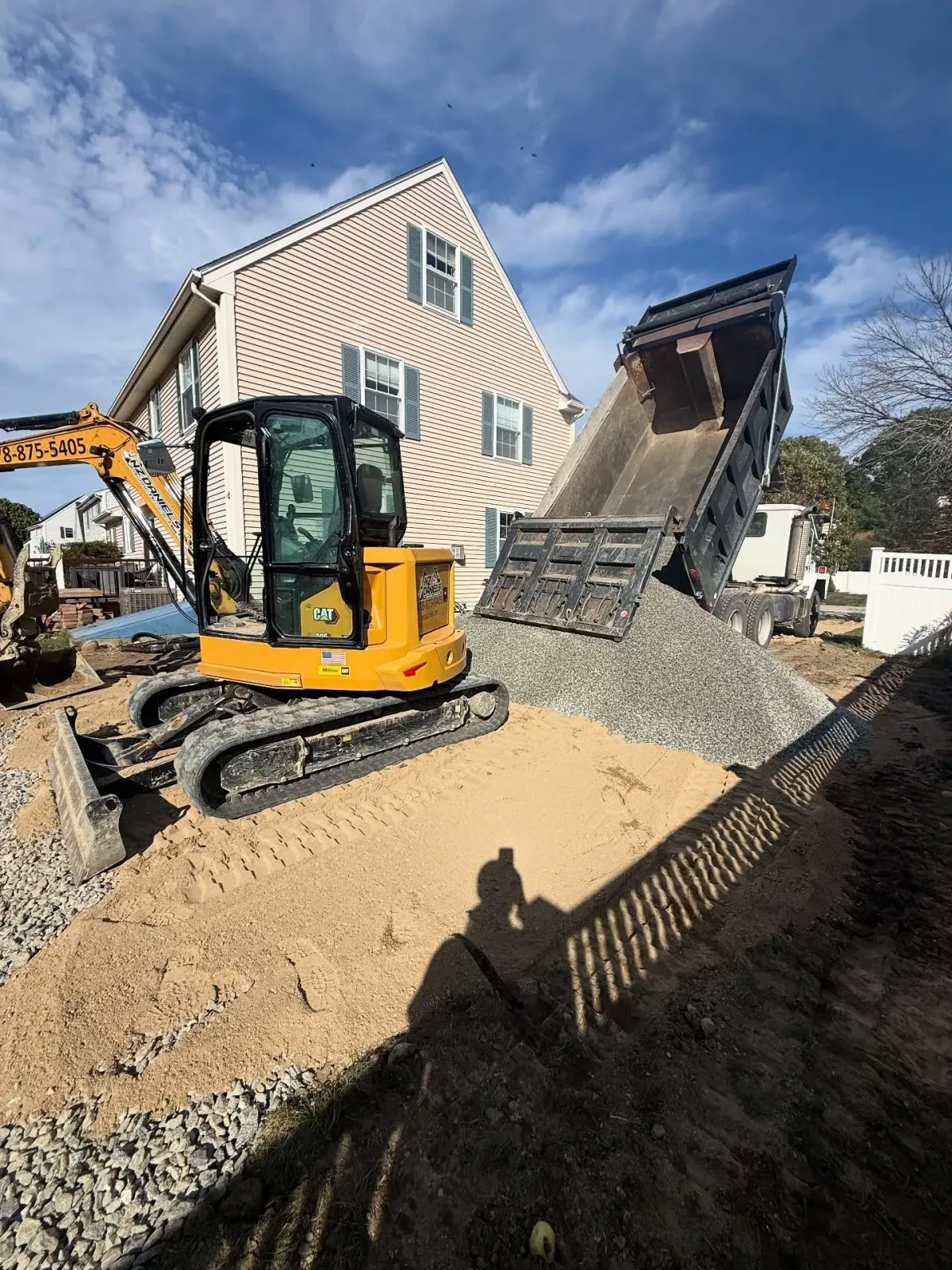 Yellow excavator next to a dump truck unloading gravel near a house. Sunny day.