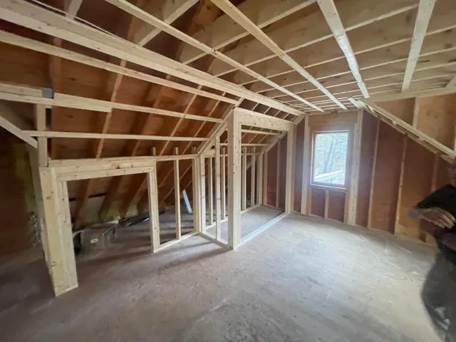 A partially constructed attic room with wooden framing, plywood flooring, and exposed beams and insulation. The room includes a window on the right side, allowing natural light to enter. Scattered construction materials and roofing siding are visible on the left.