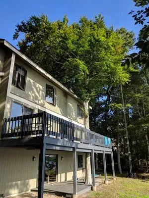 Two-story beige house with dark balcony, set among tall green trees on a sunny day