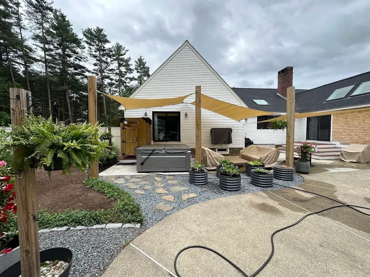 Backyard patio with a hot tub, covered furniture, and decorative planters, with a house and trees in the background under a cloudy sky.