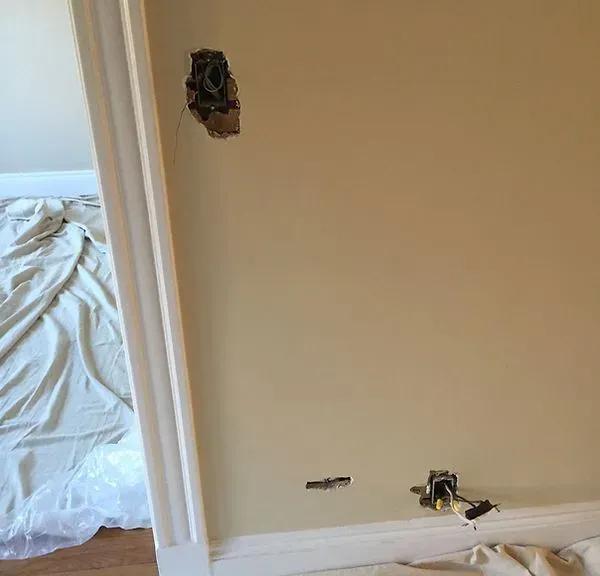 Damaged beige wall with holes and exposed wiring near a doorway in a room under repair.