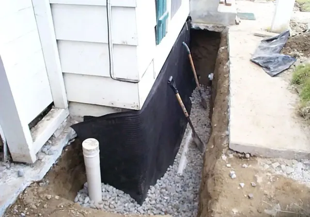 House foundation wrapped in black waterproofing with a trench and exposed pipe
