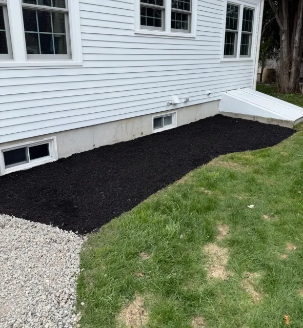 A freshly mulched garden bed next to a house with white siding, small window wells, and a small basement window; the garden bed is edged with white gravel and bordered by a grassy lawn.