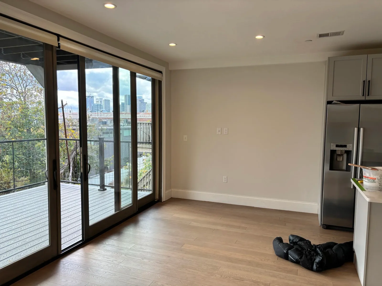 Empty modern living room with wooden floor, sliding glass doors leading to a balcony with a city view, and a stainless steel refrigerator to the right.