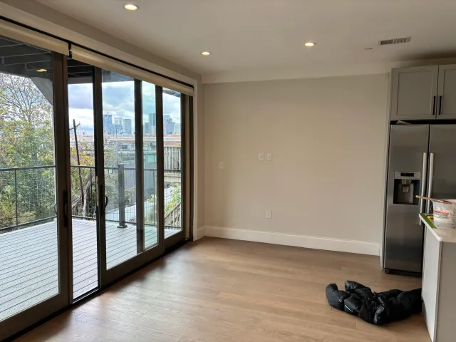 Empty modern living room with wooden floor, sliding glass doors leading to a balcony with a city view, and a stainless steel refrigerator to the right.