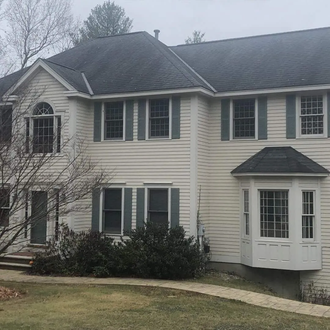 Two-story house with gray siding and shutters, large windows, and a bay window extension. The house is surrounded by a small lawn with shrubs and a bare tree. Overcast sky in the background.
