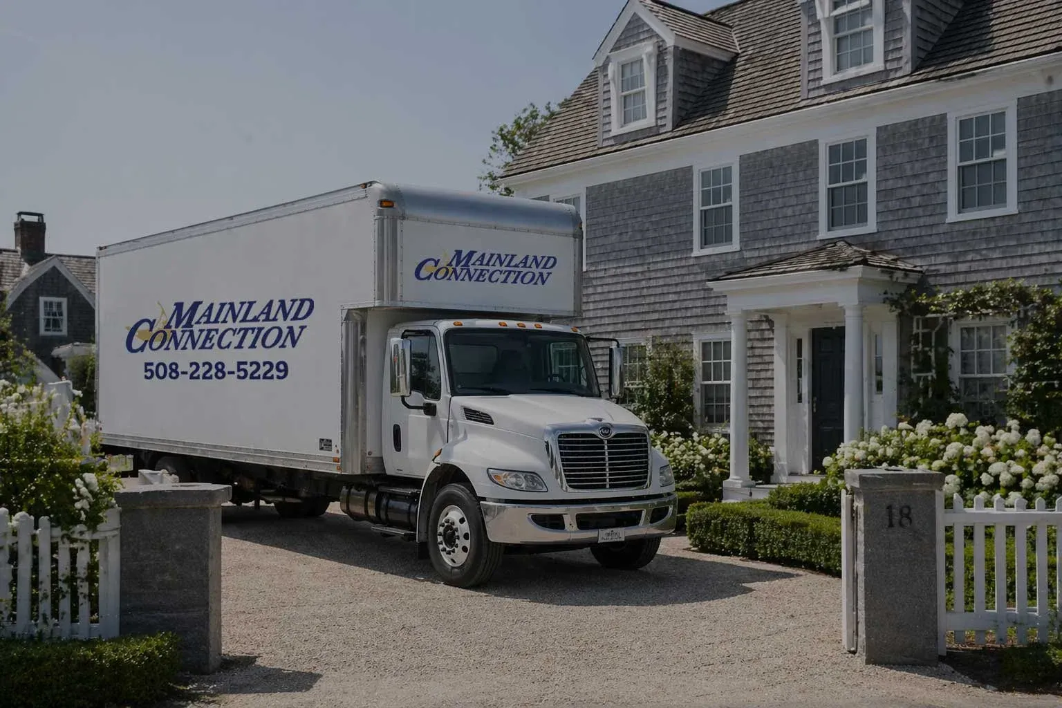 White Nantucket moving truck with 'Mainland Connection' branding parked in front of a house with gray siding and a porch, surrounded by flowering bushes and a white picket fence.