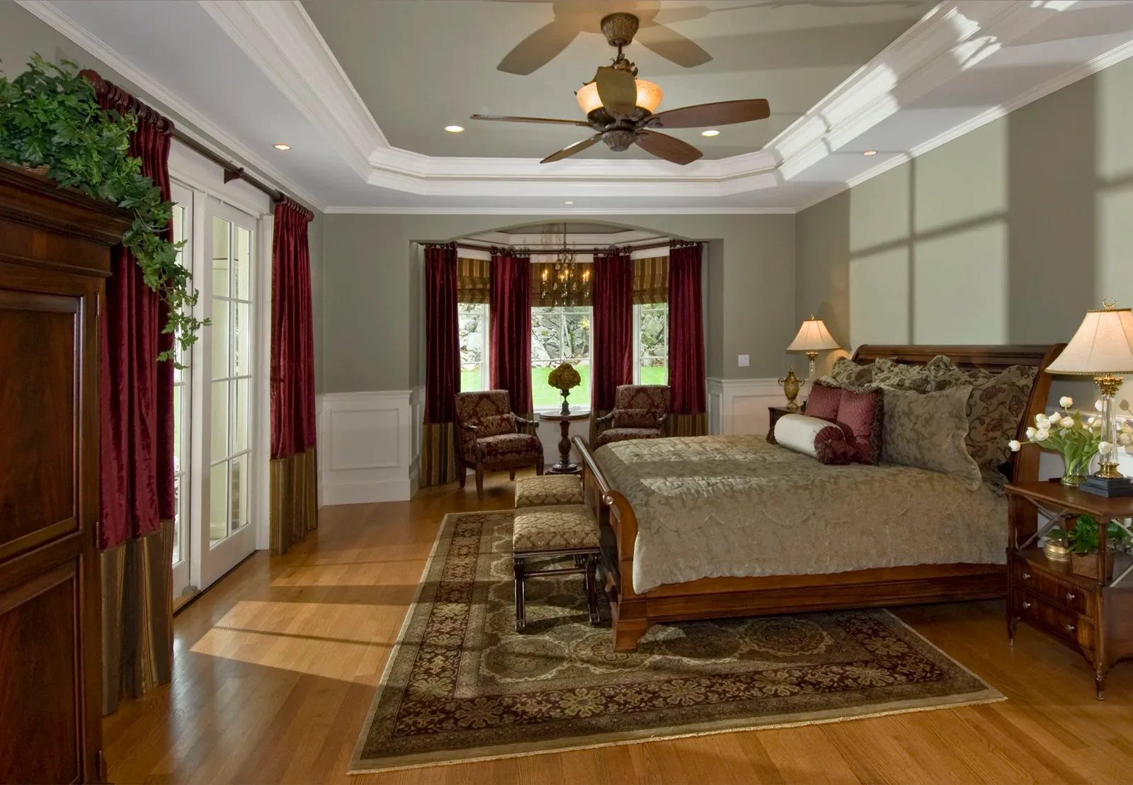 A spacious bedroom with a large bed, wooden furniture, and a seating area near a window with red curtains. The room has hardwood flooring, a decorative area rug, and a ceiling fan.