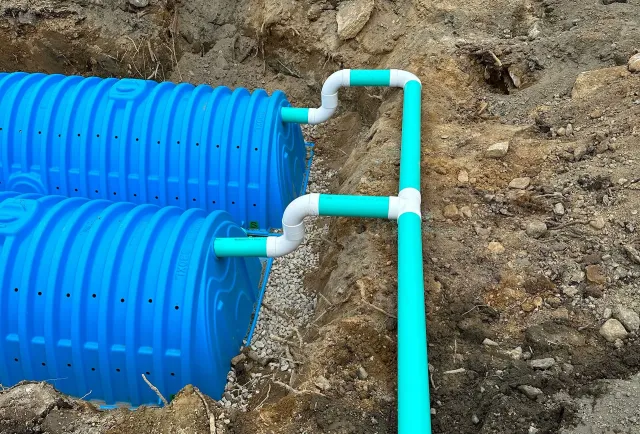 Two blue plastic septic chambers installed in a trench, connected by white pipes to a central green distribution line.