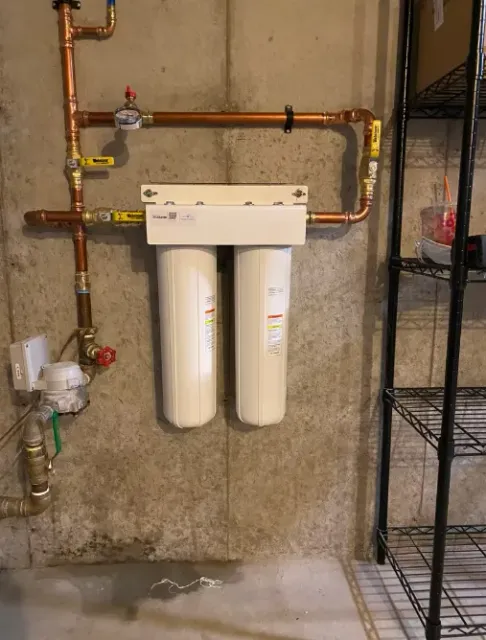 Wall-mounted water filtration system with pipes, two white filter canisters, and shelving in a utility room