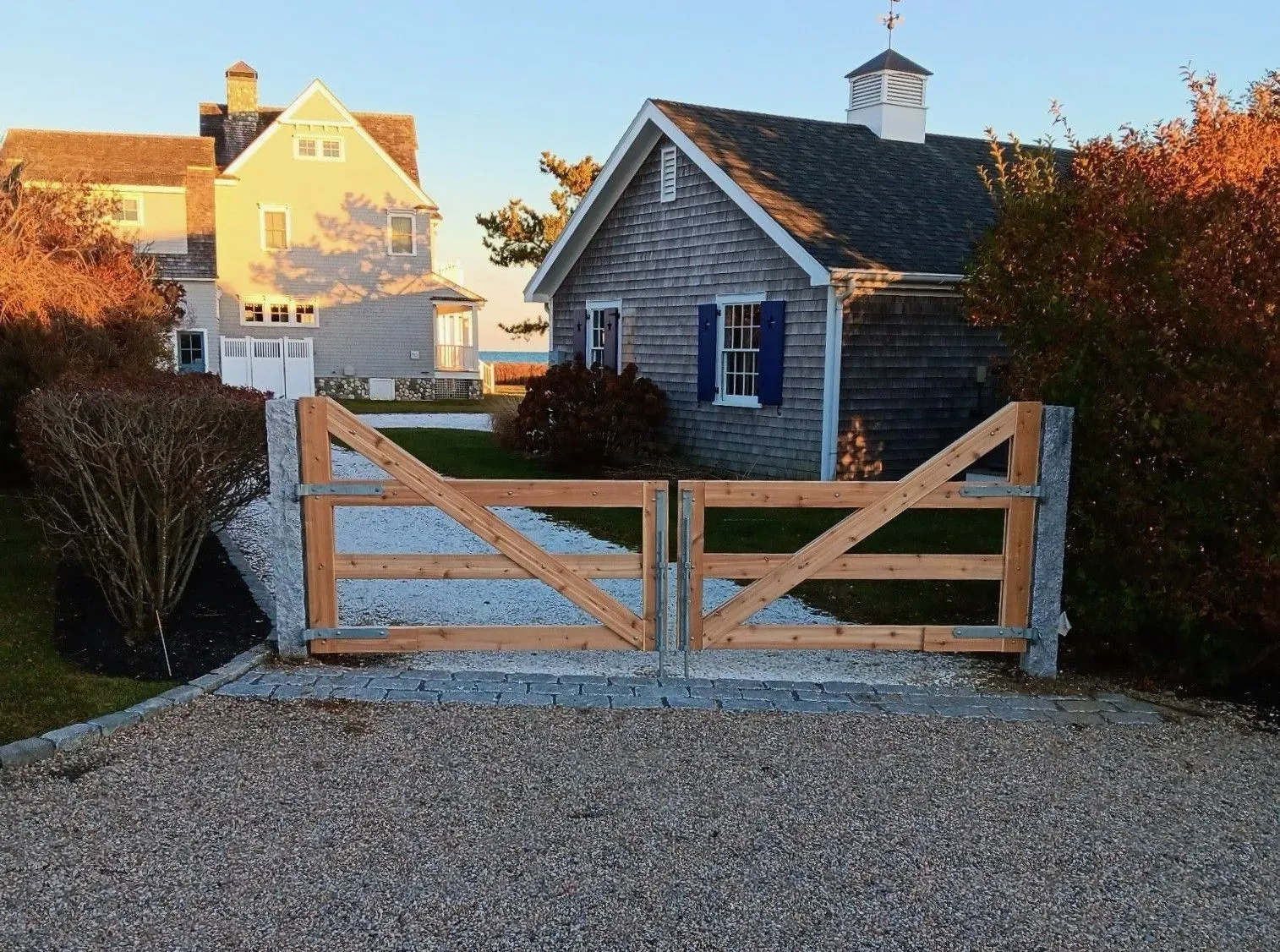 A wooden gate stands at the entrance of a gravel driveway between a cedar-shingled cottage and a larger pale house.