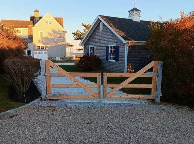 A wooden gate stands at the entrance of a gravel driveway between a cedar-shingled cottage and a larger pale house.
