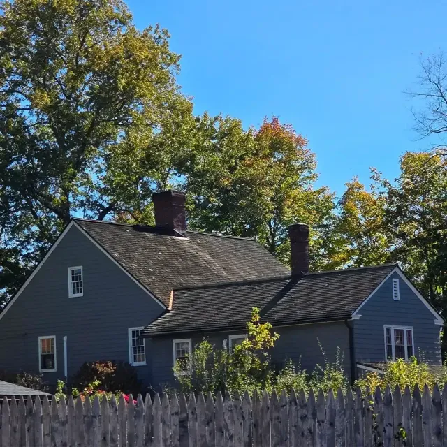 A gray, traditional-style house with two chimneys and a dark shingled roof, set against trees under a clear blue sky.