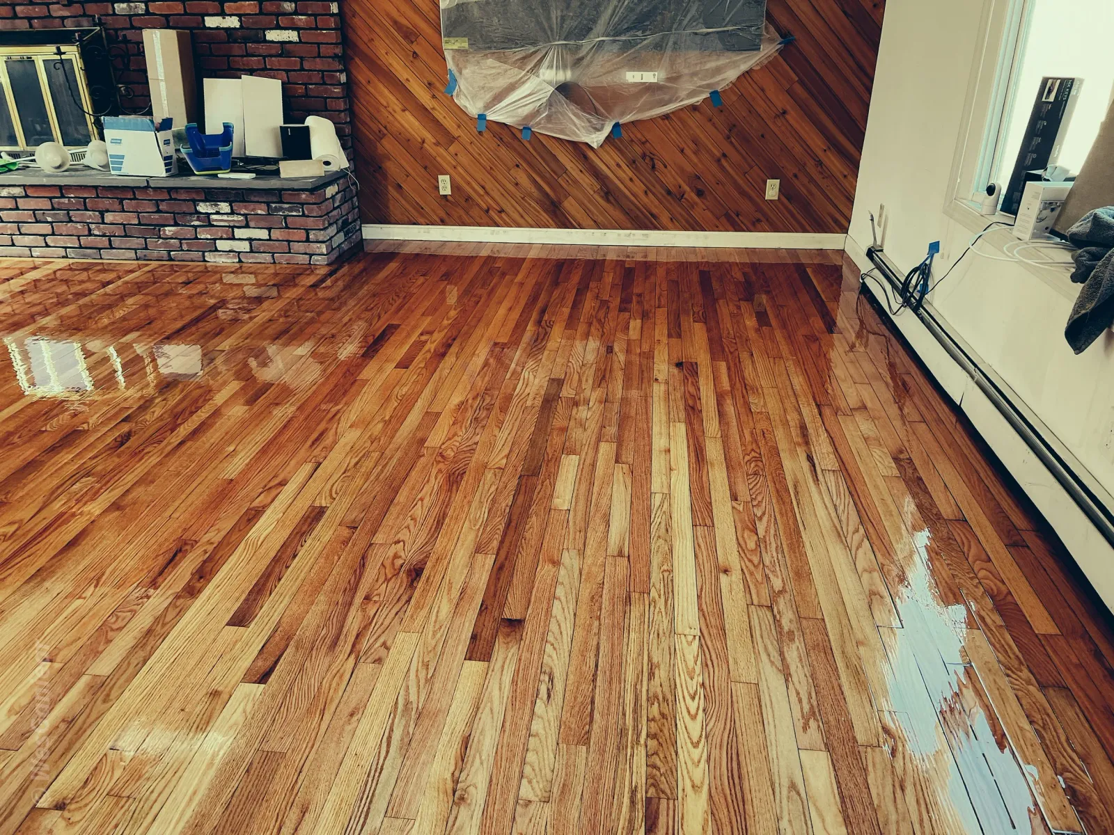 Refinished hardwood floor in a room, with some areas still wet and drying, and an empty space near the window with tools and materials on the brick fireplace ledge.