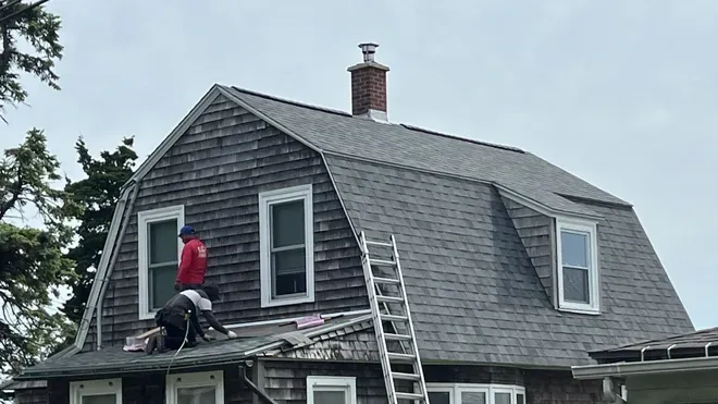 Worker on a ladder repairing a gray shingled roof on a house