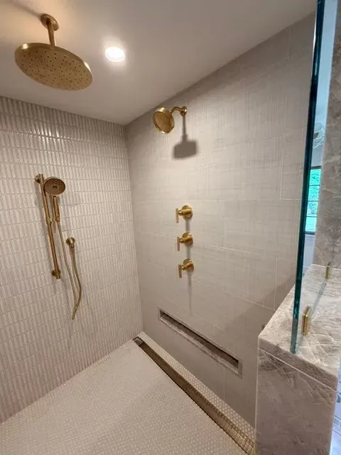 Modern shower with gold fixtures, tiled walls, bench, and marble accent ledge.
