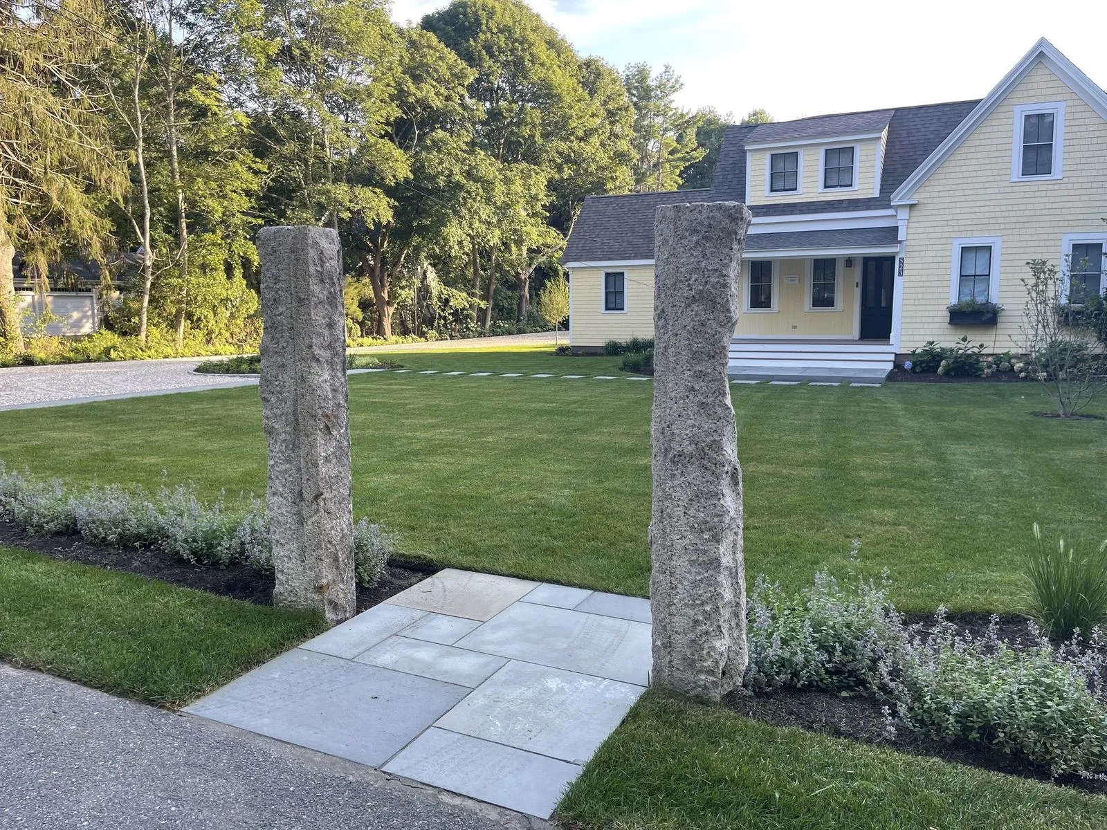 Two granite monolith pillars frame a bluestone walkway leading to the front door of a yellow Cape Cod home with a manicured front lawn