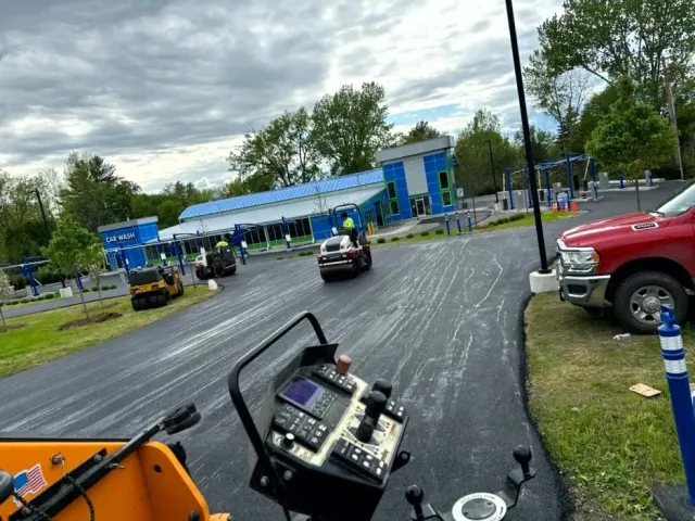 Asphalt paving at a blue car wash. Vehicles and workers visible. Cloudy sky.