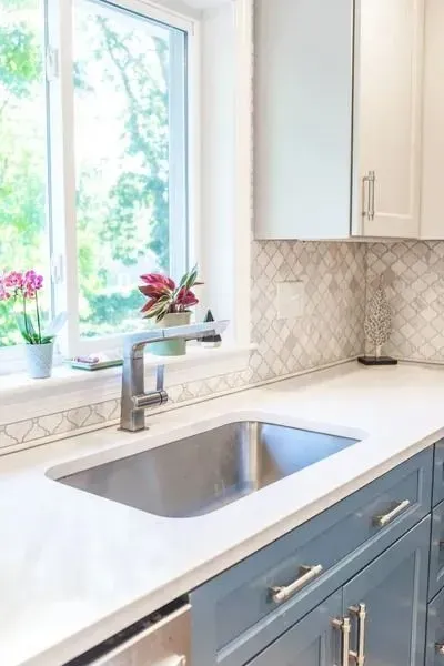Bright kitchen sink by a window, with white counters and blue lower cabinets.