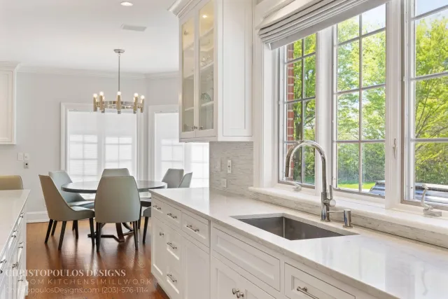 Bright kitchen with white cabinets, marble countertops, a large window with a view of green trees, and a dining area with beige chairs.