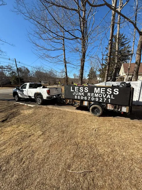 White truck towing a Less Mess Junk Removal trailer on brown grass, trees in the background, sunny day.