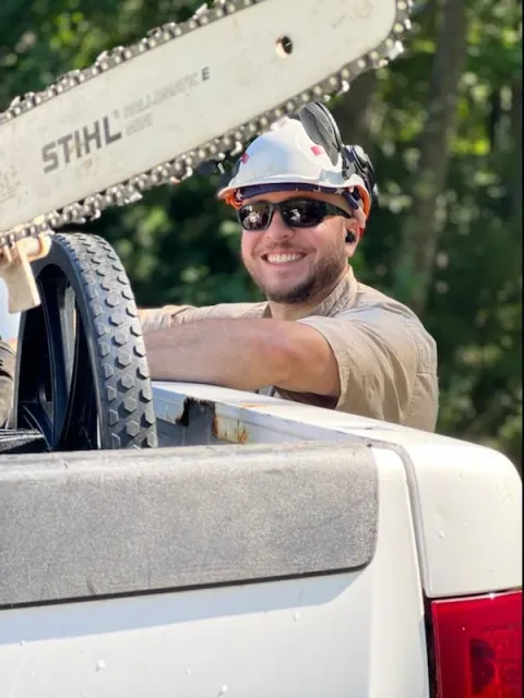 A smiling man wearing sunglasses, a beige shirt, and a white hard hat with earmuffs, standing in the bed of a pickup truck with a chainsaw in the foreground, outdoors with trees in the background.