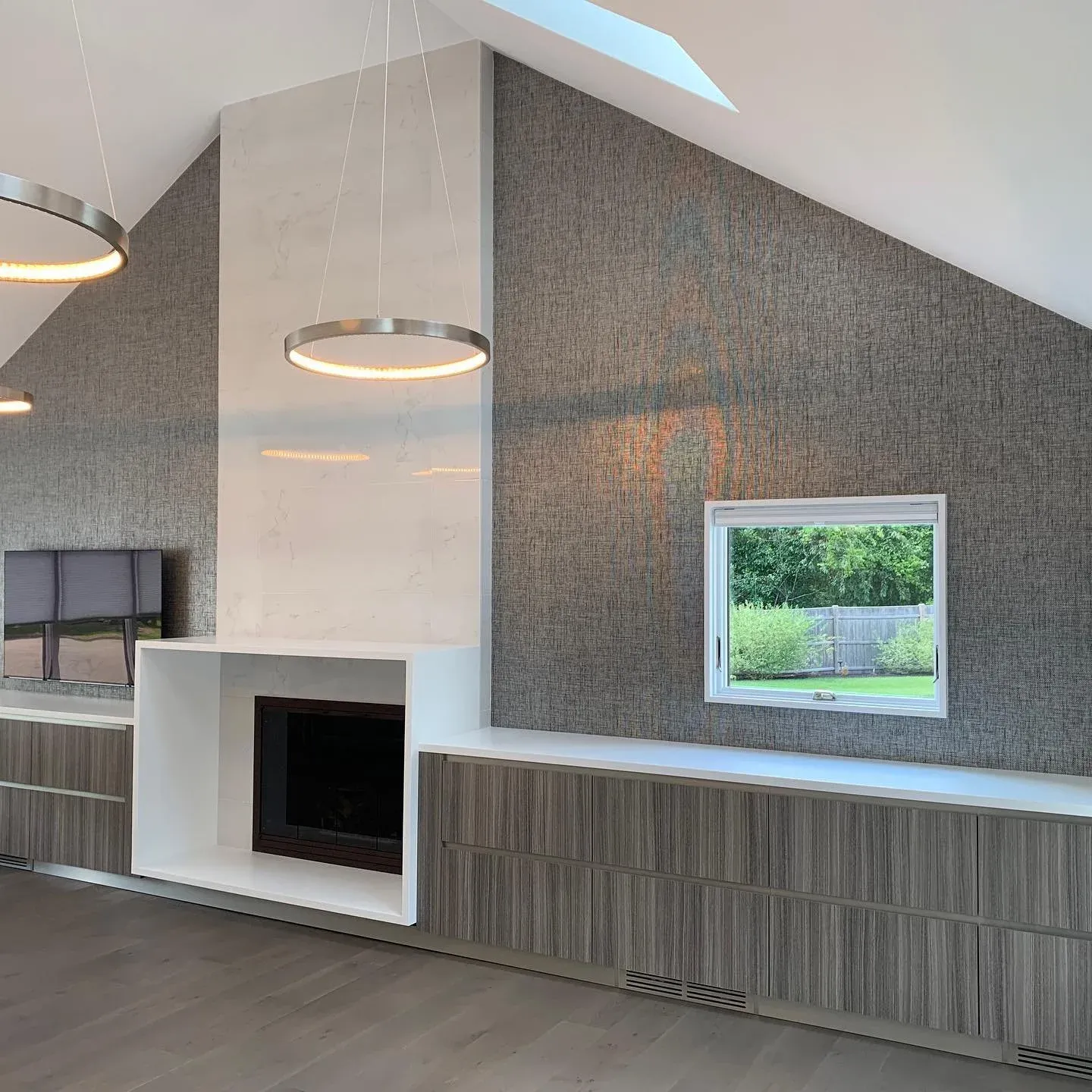 Modern living room with built-in white fireplace, textured gray wall, and large wall-mounted TV. Ceiling with skylight and circular pendant lights.