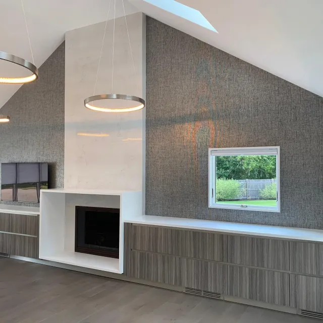 Modern living room with built-in white fireplace, textured gray wall, and large wall-mounted TV. Ceiling with skylight and circular pendant lights.