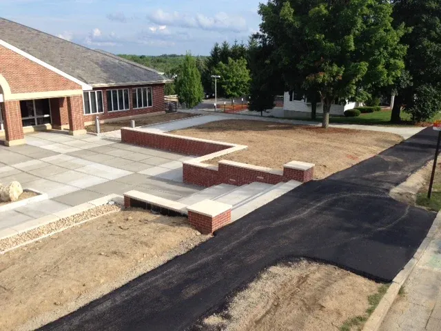 A sidewalk and driveway outside a building during daytime, with a small brick wall, trees, and a partly cloudy sky.