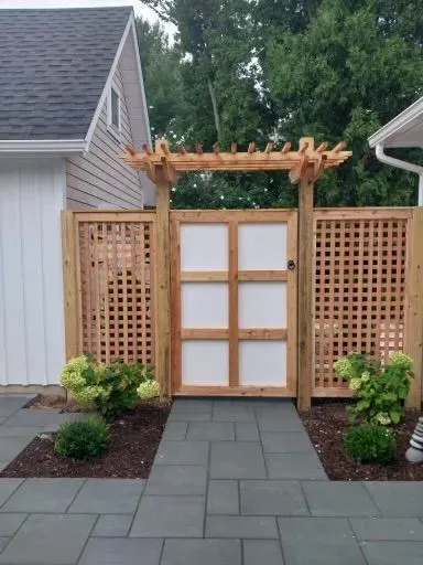 A wooden fence with a gate and a pergola in front of a house.