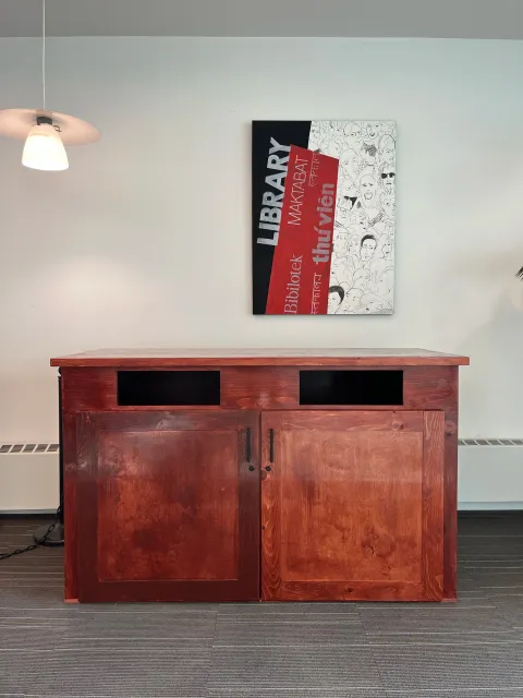 Wooden cabinet with closed doors on a dark gray carpeted floor, above it a colorful poster on the white wall. To the left, part of a hanging lamp is visible.