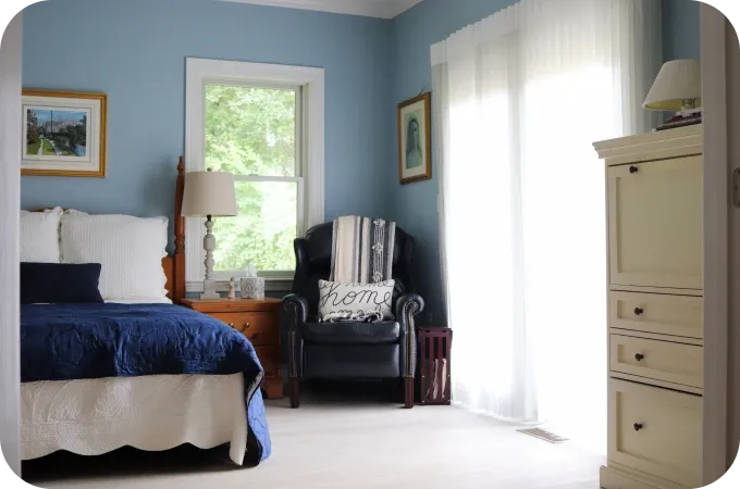 Blue bedroom with white bed, navy quilt, nightstand, lamp, black armchair, and dresser.