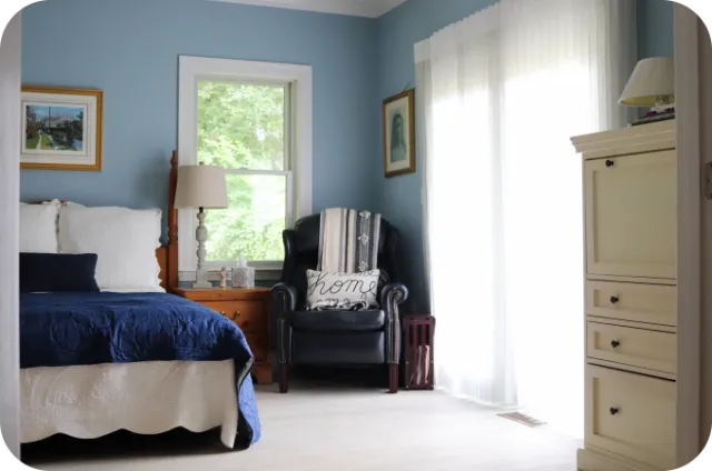 Blue bedroom with white bed, navy quilt, nightstand, lamp, black armchair, and dresser.