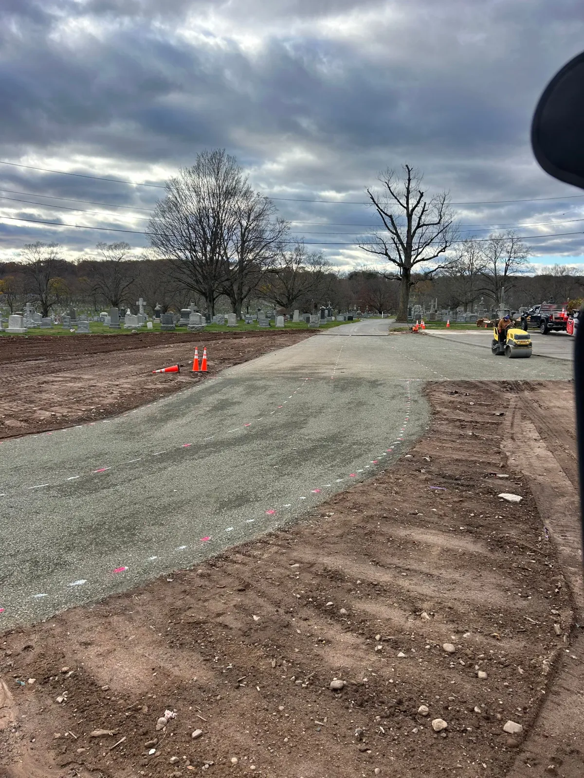A dirt road leads through a cemetery with bare trees under a cloudy sky, with construction equipment visible in the distance.