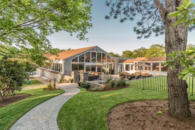 A spacious backyard with a landscaped lawn, a stone pathway, and a multi-section house with large windows, a patio area with outdoor furniture, umbrellas, and a fire pit, surrounded by trees and greenery.