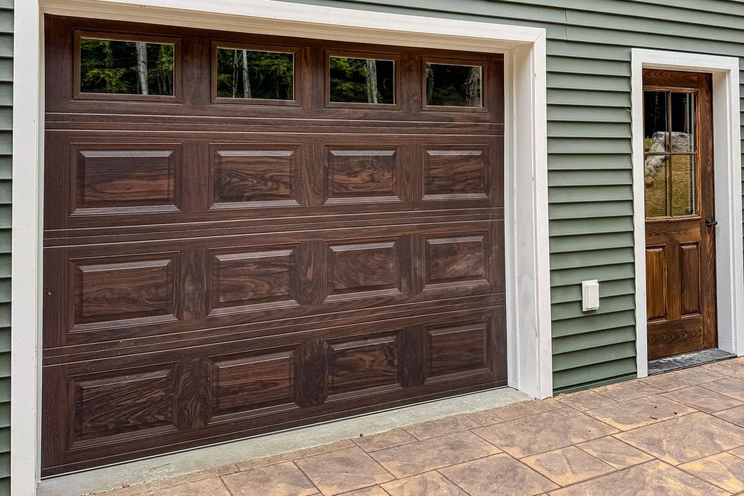 Garage door with windows next to a wooden door on a green siding house.