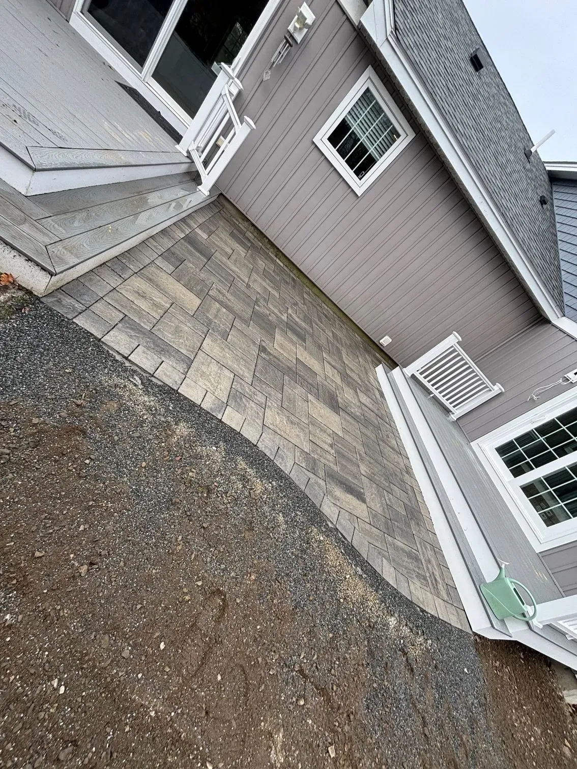 Paver patio beside a light gray house. Gravel path and exposed soil in foreground.